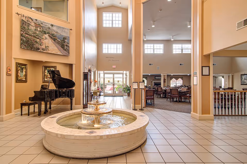 Spacious and well-lit interior of Creekside Pines Retirement Community featuring a central round water fountain, a grand piano to the left, and a dining area with tables and chairs in the background. The area has high ceilings with large windows allowing natural light to fill the space. Artwork is displayed on the walls, and the floor is tiled.