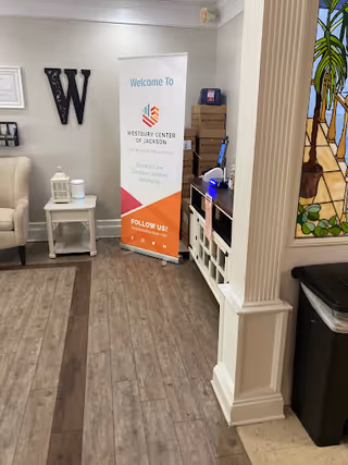 Interior view of a senior living facility reception or common area with a welcome banner for Westbury Center of Jackson for Nursing and Healing, a beige armchair, a small white side table with a lantern and a diffuser, wooden flooring, and a stained glass window on the right side.