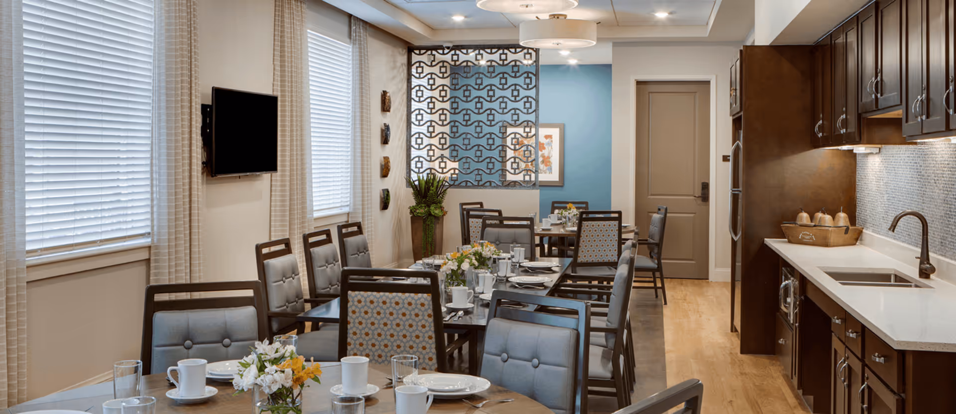 Communal dining room set for a meal with multiple tables and chairs, a wall-mounted TV, decorative screen, and a kitchenette with sink and cabinets.