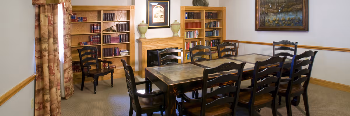 A cozy room with a large wooden table surrounded by eight dark wooden chairs. The room features two wooden bookshelves filled with books, a fireplace mantle with decorative vases, floral patterned curtains on the window, and a framed painting on the wall.