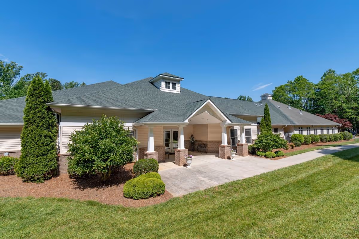 Exterior view of a single-story senior living facility building with beige siding, a green roof, and a covered entrance supported by white columns. The building is surrounded by well-maintained landscaping including bushes, small trees, and a green lawn under a clear blue sky.