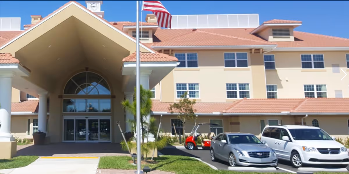 Front exterior of a retirement community showing the main entrance with a covered porte-cochere, an American flag, and parked vehicles.