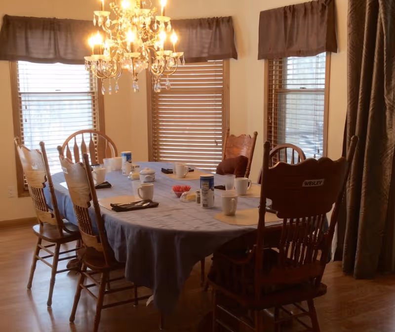 Dining room with a cloth-covered rectangular table set with mugs and condiments, surrounded by wooden chairs under a chandelier by windows with blinds.