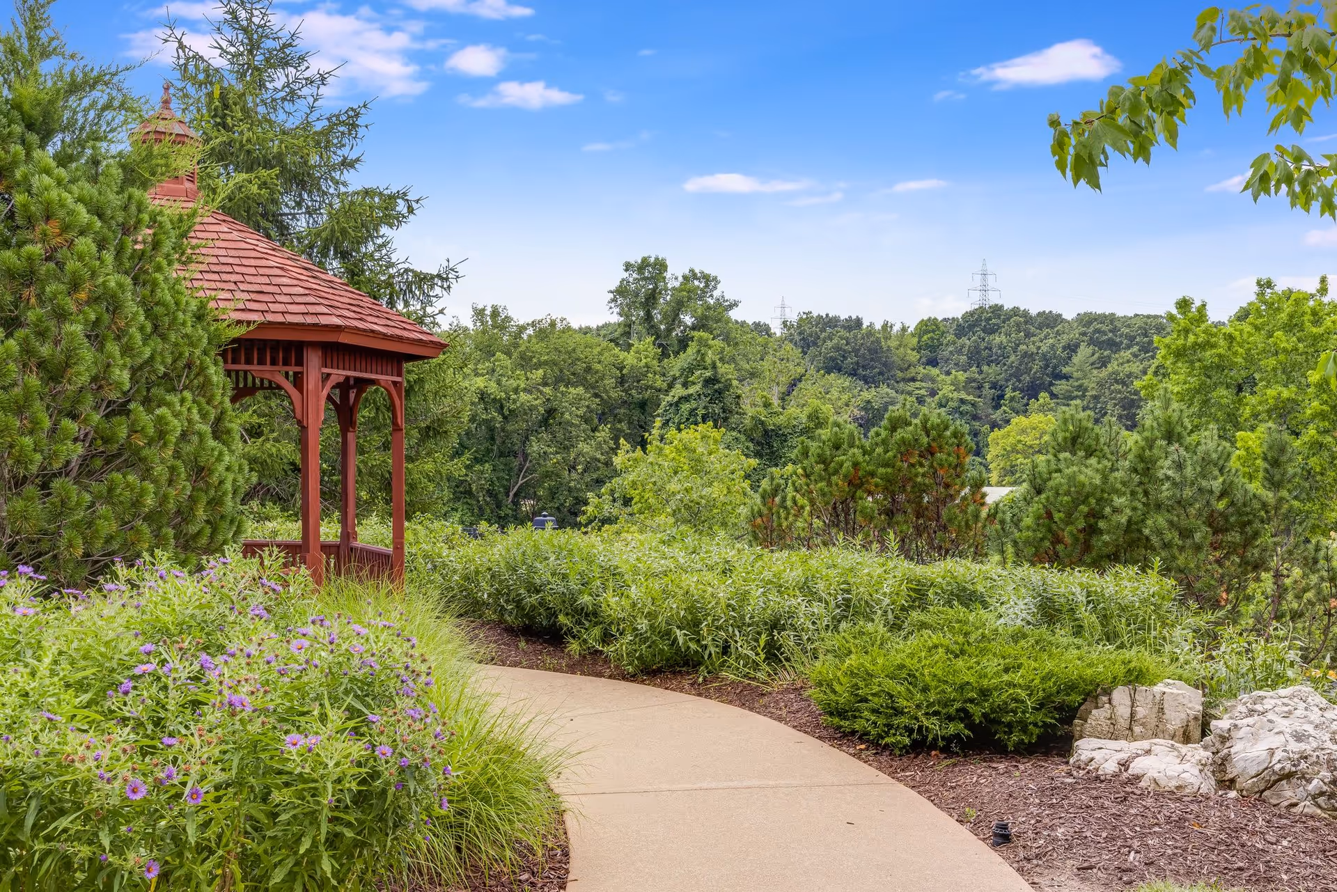 A winding concrete pathway through a lush garden with various green shrubs, purple flowers, and trees. To the left, there is a red wooden gazebo with a shingled roof. The sky is clear with a few clouds.