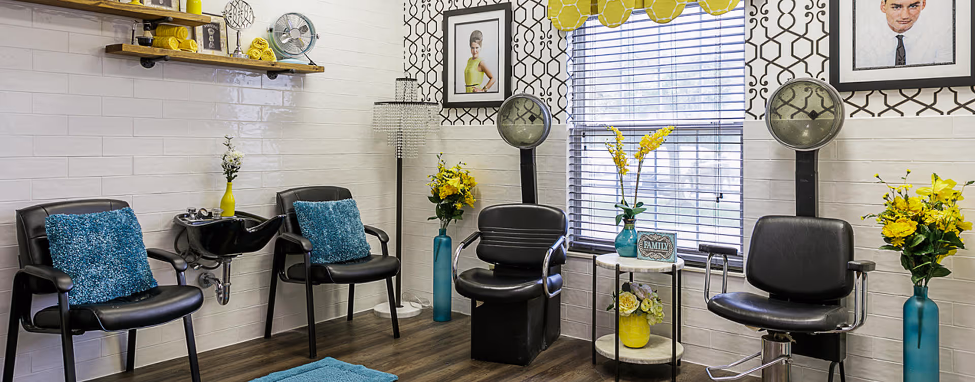 Interior of a hair salon area with four black salon chairs, two hair washing sinks, a small round table with flowers and a family sign, and decorative framed pictures on the walls. The room has white tiled walls, wooden flooring, and a window with blinds letting in natural light.