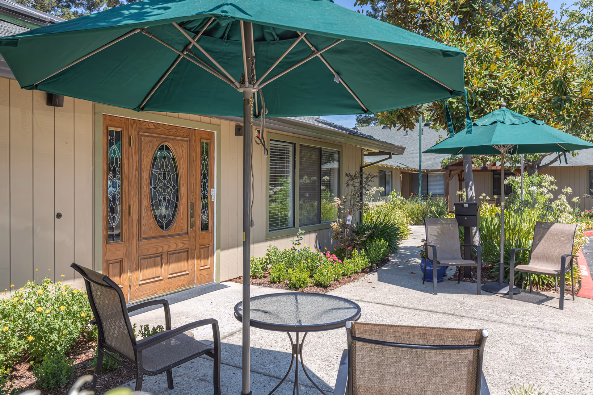 Outdoor patio area at Almond Grove Assisted Living with green umbrellas shading glass-top tables and mesh chairs. The patio is adjacent to a building with a wooden door featuring decorative glass panels and windows with blinds. Surrounding the patio are green plants and flowers under a clear blue sky.
