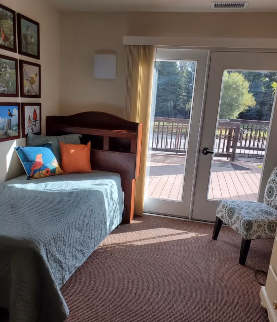 Sunlit small bedroom with a single bed, colorful pillows and bird artwork, and glass doors opening onto a deck.