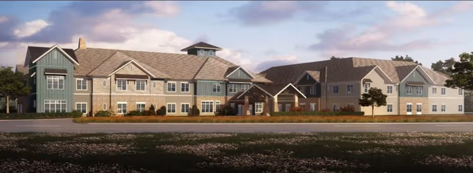 Exterior view of a large two-story senior living facility building with a combination of green and beige siding, multiple windows, and a covered entrance. The building is surrounded by landscaped grounds and a road in the foreground under a partly cloudy sky.