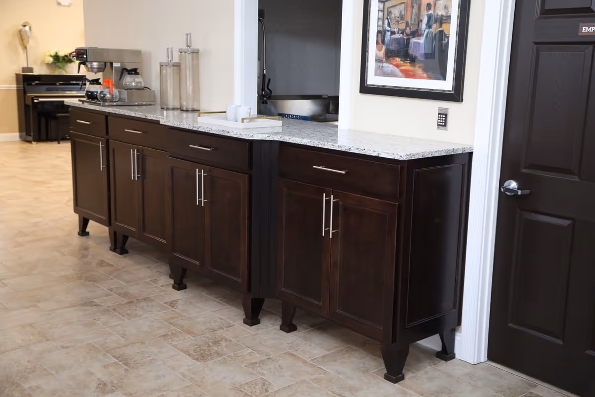 A hallway area with dark wood cabinets topped with a light granite countertop. On the countertop, there is a coffee maker, coffee pots, and two glass containers. In the background, there is a piano and a beige wall with a small plant on top of the piano. A framed painting hangs on the wall above the cabinets, and a dark door is visible to the right.