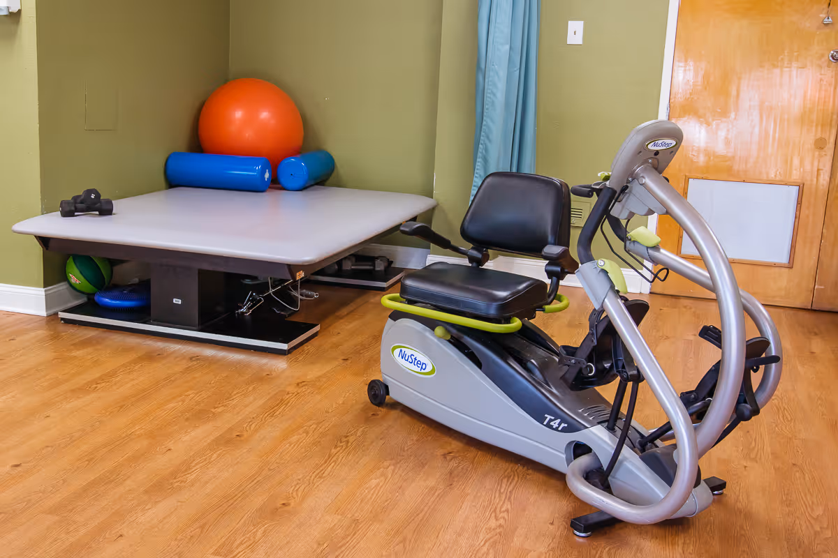 A small exercise room with a NuStep T4r recumbent cross trainer, a padded therapy table with various exercise equipment including an orange exercise ball, blue foam rollers, and small dumbbells. The room has wooden flooring, green walls, and a wooden door.