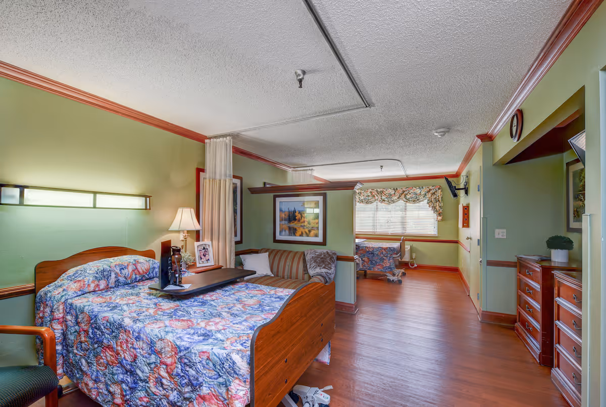 A senior living facility room with a wooden bed covered in a colorful floral quilt. Next to the bed is a small table with a lamp, a photo frame, and a tray. The room has green walls with wooden trim and a wooden floor. There is a small sofa with pillows and a blanket, a window with floral curtains, a wall-mounted TV, and a wooden dresser with a small plant on top.