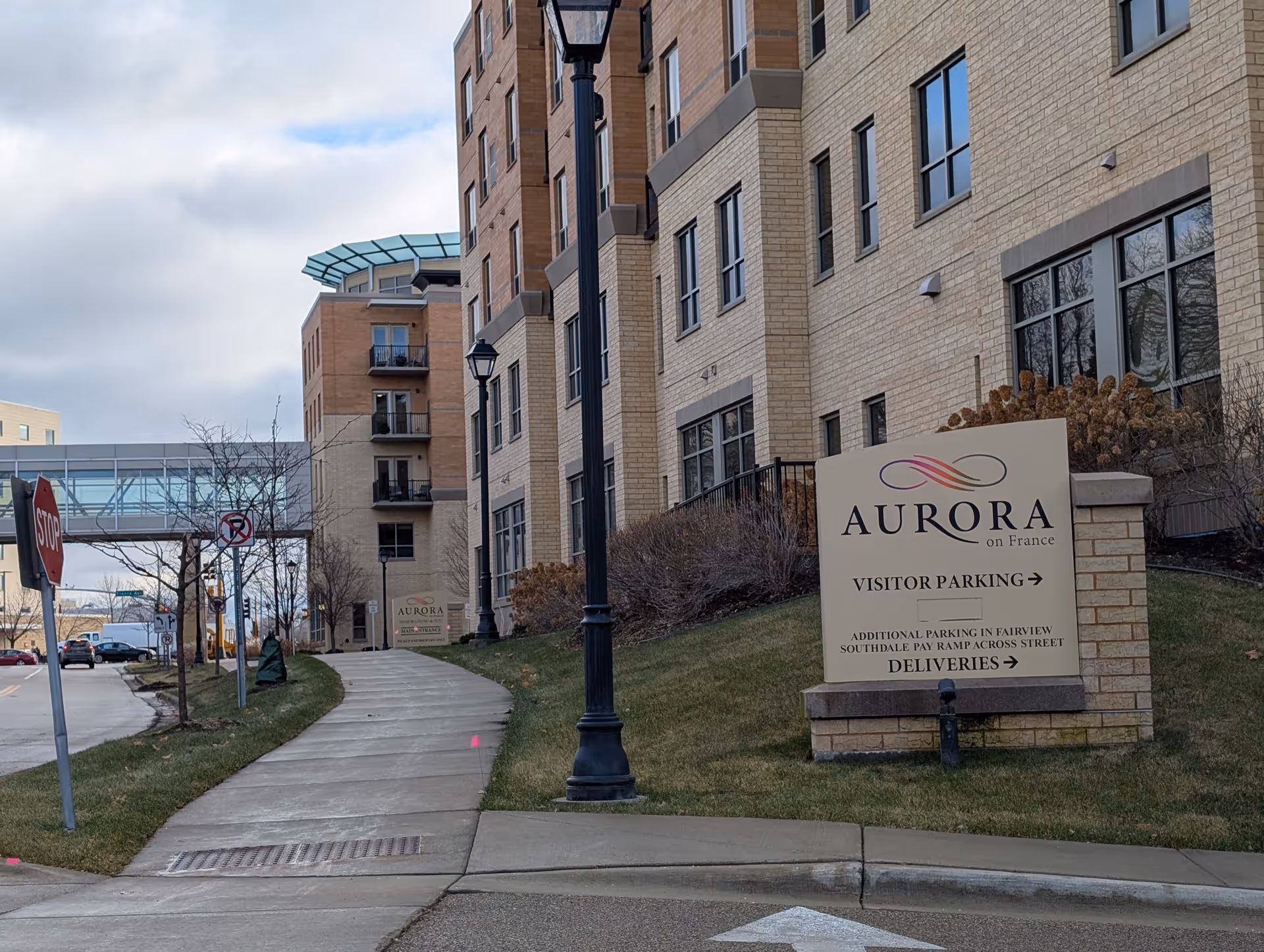 Exterior view of the Aurora on France senior living facility showing a sidewalk leading to the entrance, a building with multiple windows, a street lamp, and a sign indicating visitor parking and deliveries.