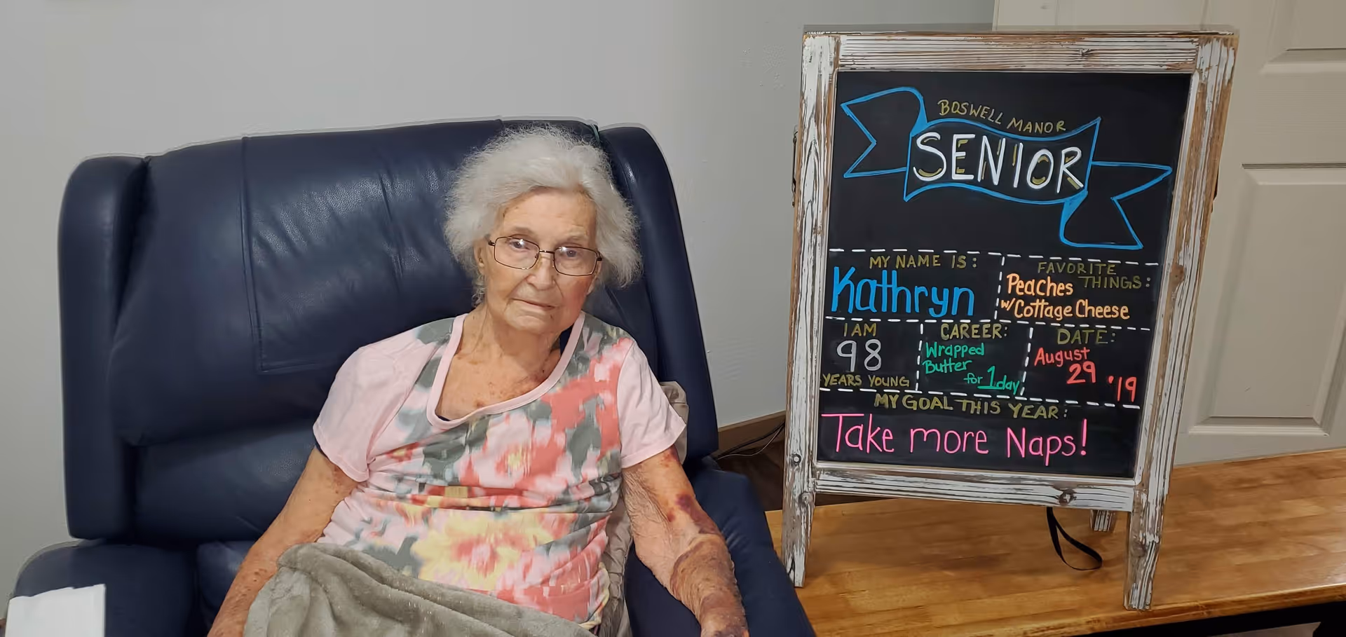 An elderly woman with white hair and glasses sitting in a dark blue recliner chair. She is wearing a floral patterned shirt and has a blanket over her lap. Next to her is a chalkboard sign with colorful writing that reads: Boswell Manor Senior. My name is Kathryn. I am 98 years young. Career: Wrapped Butter for 1 day. Favorite things: Peaches w/ Cottage Cheese. Date: August 29 '19. My goal this year: Take more naps!