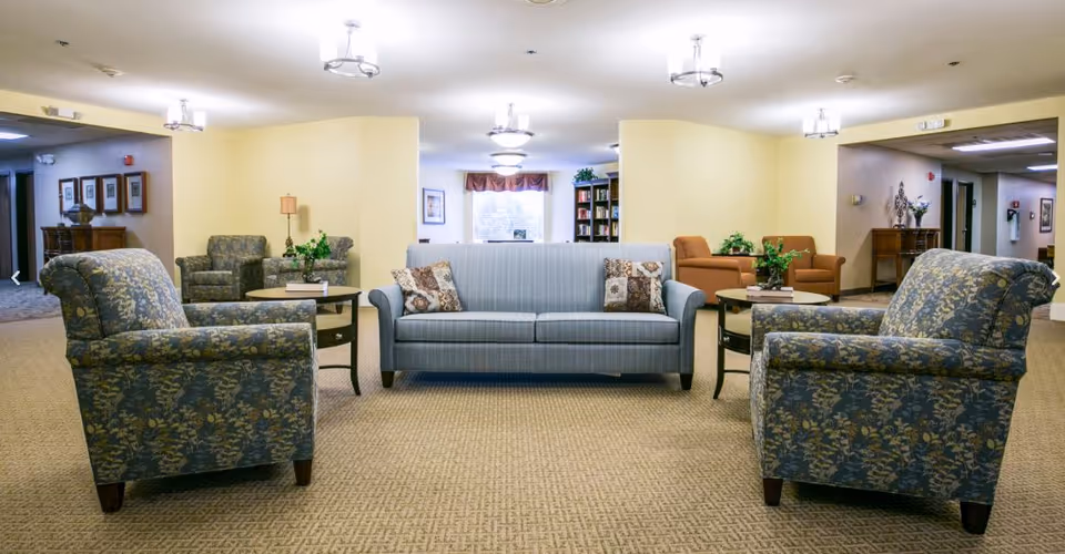 A well-lit common area in a senior living facility featuring a blue striped sofa with two patterned pillows, two floral upholstered armchairs facing the sofa, and two round wooden side tables with plants and books. In the background, there are additional seating areas with armchairs, a bookshelf, framed pictures on the walls, and ceiling lights.