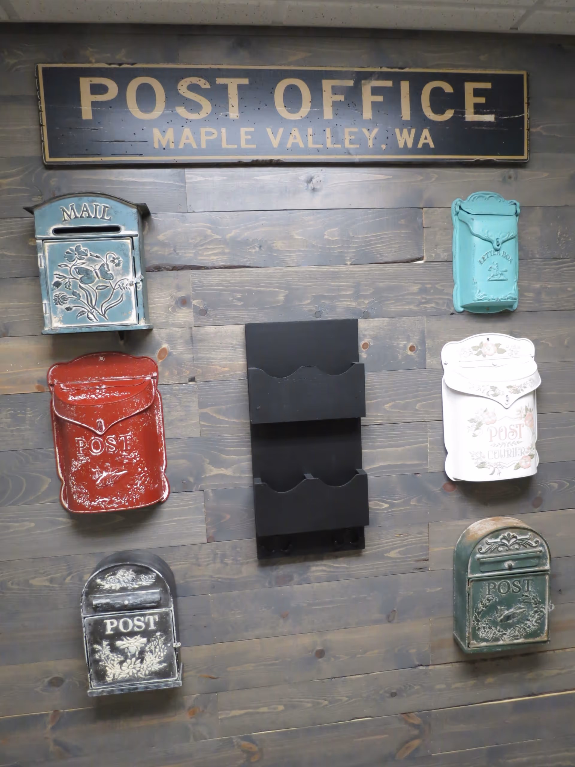 A rustic wooden wall with a vintage-style sign reading 'POST OFFICE MAPLE VALLEY, WA' at the top. Below the sign are six decorative mailboxes in various colors and designs, including blue, red, black, white, turquoise, and green. There is also a black double-slot organizer mounted in the center.