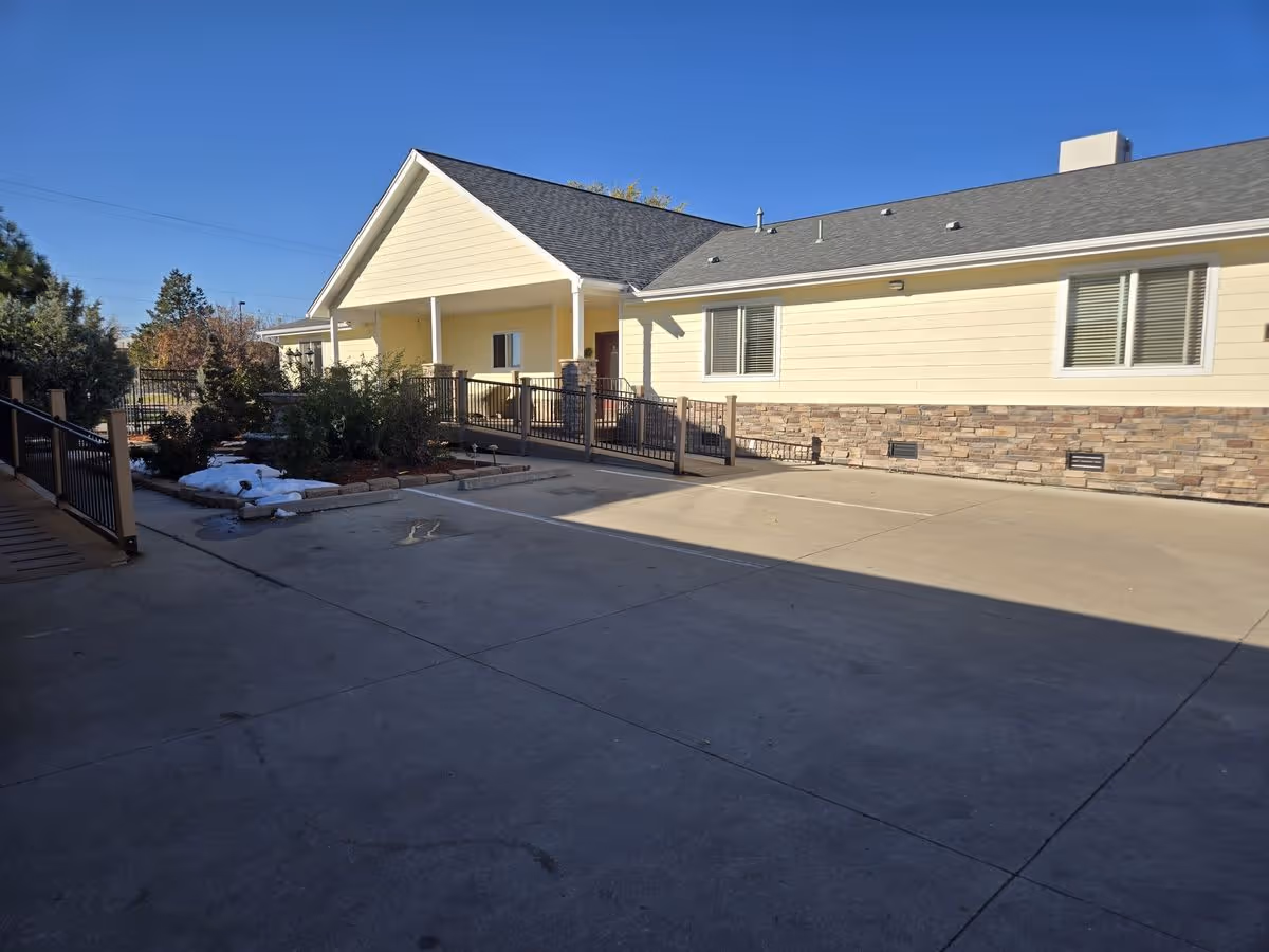 Exterior courtyard and entrance of a single-story light-yellow building with a ramp, railing, and small landscaped area.