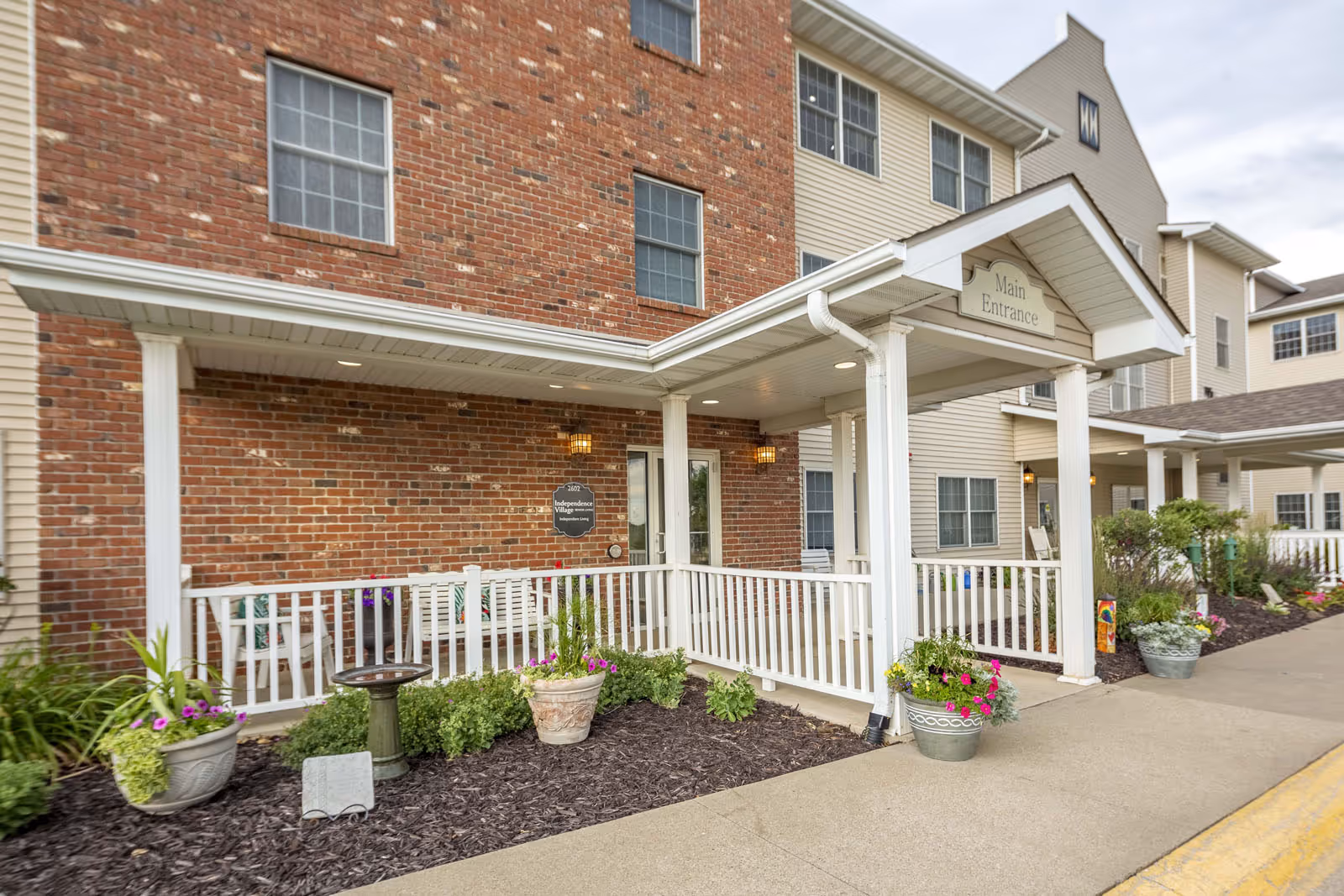 Covered main entrance of a multi-story brick-and-siding senior living building with white porch railings, potted flowers, and a 'Main Entrance' sign.