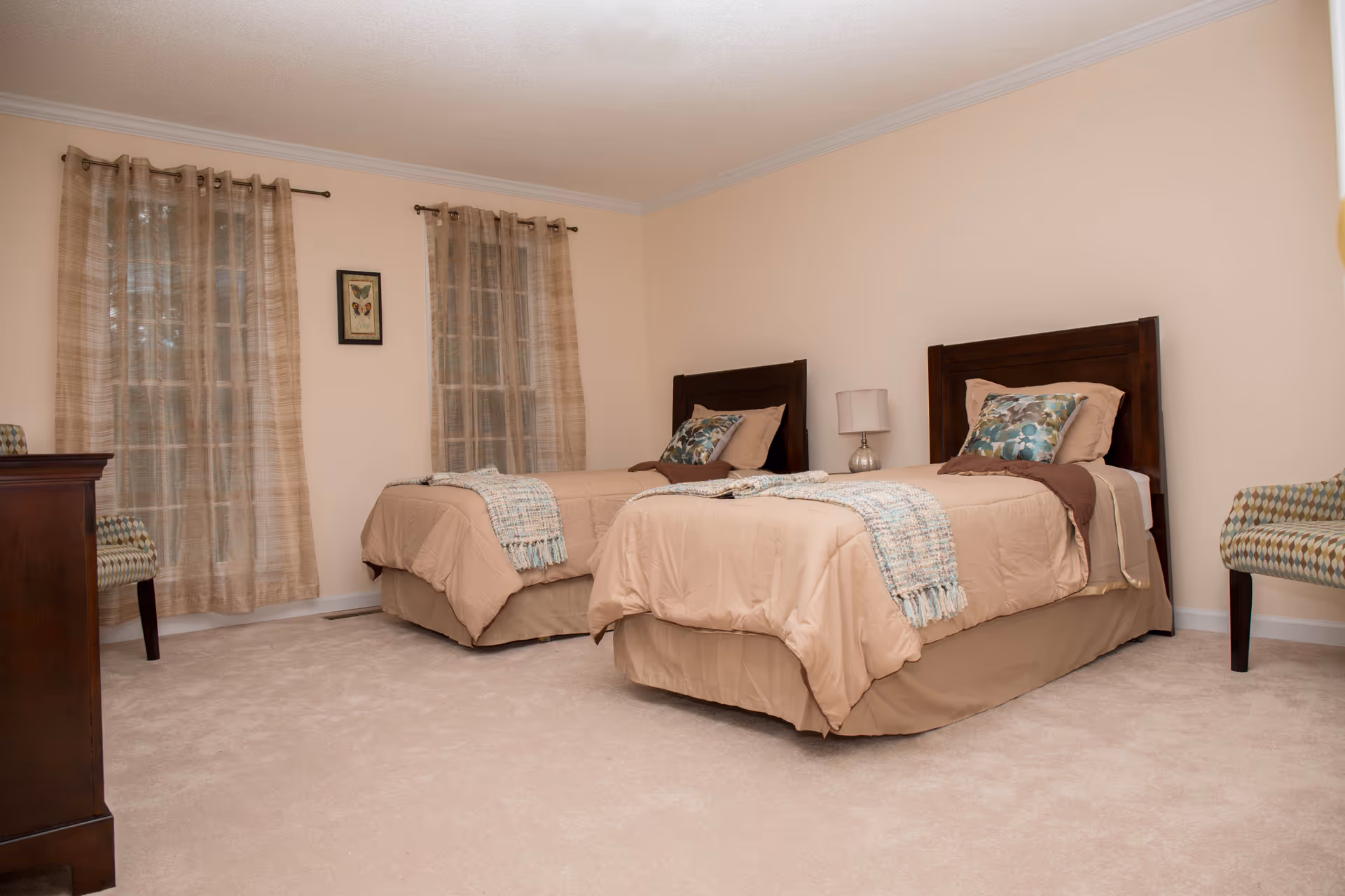A bedroom with two twin beds featuring beige bedding and decorative pillows. The room has beige walls, two windows with sheer beige curtains, a small lamp on a nightstand between the beds, a wooden dresser, and two patterned chairs.
