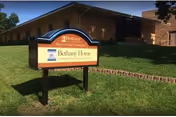 Outdoor view of Bethany Home Sioux Falls building with a sign in the foreground on a grassy area. The sign displays the facility name and tagline, with the building and trees visible in the background under a clear sky.