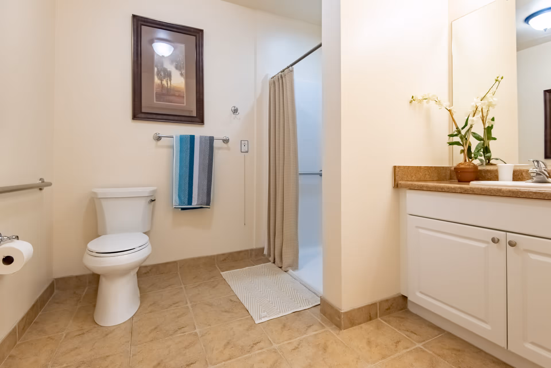 A clean bathroom with beige tiled floor and walls painted in light cream. The room features a white toilet, a towel rack with a blue and white striped towel, a walk-in shower with a beige curtain, a white vanity with a brown countertop, a sink, and a potted plant with white flowers. A framed picture hangs above the towel rack.