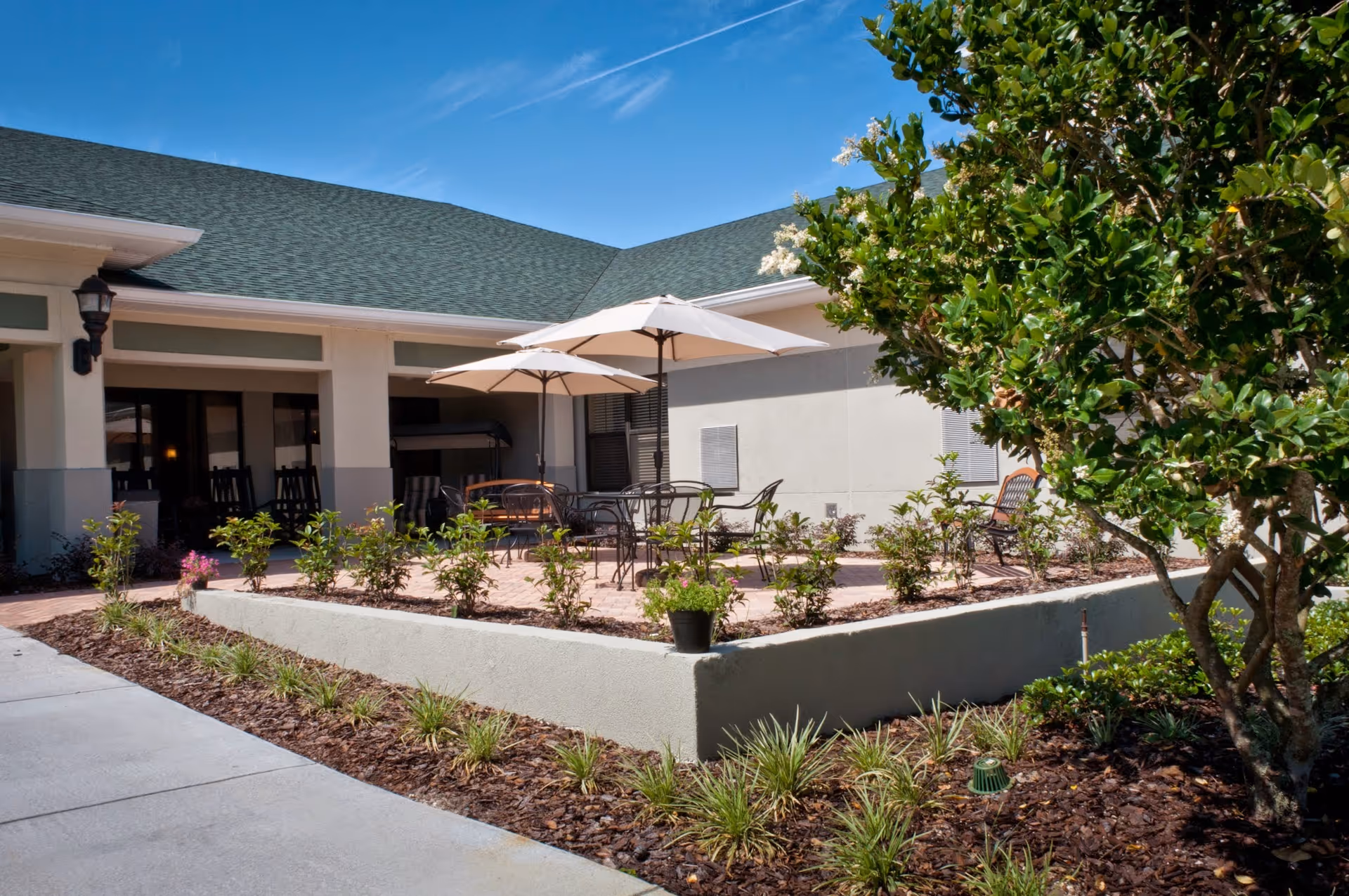 Outdoor patio area at a senior living facility with tables, chairs, and umbrellas surrounded by plants and landscaping under a clear blue sky.