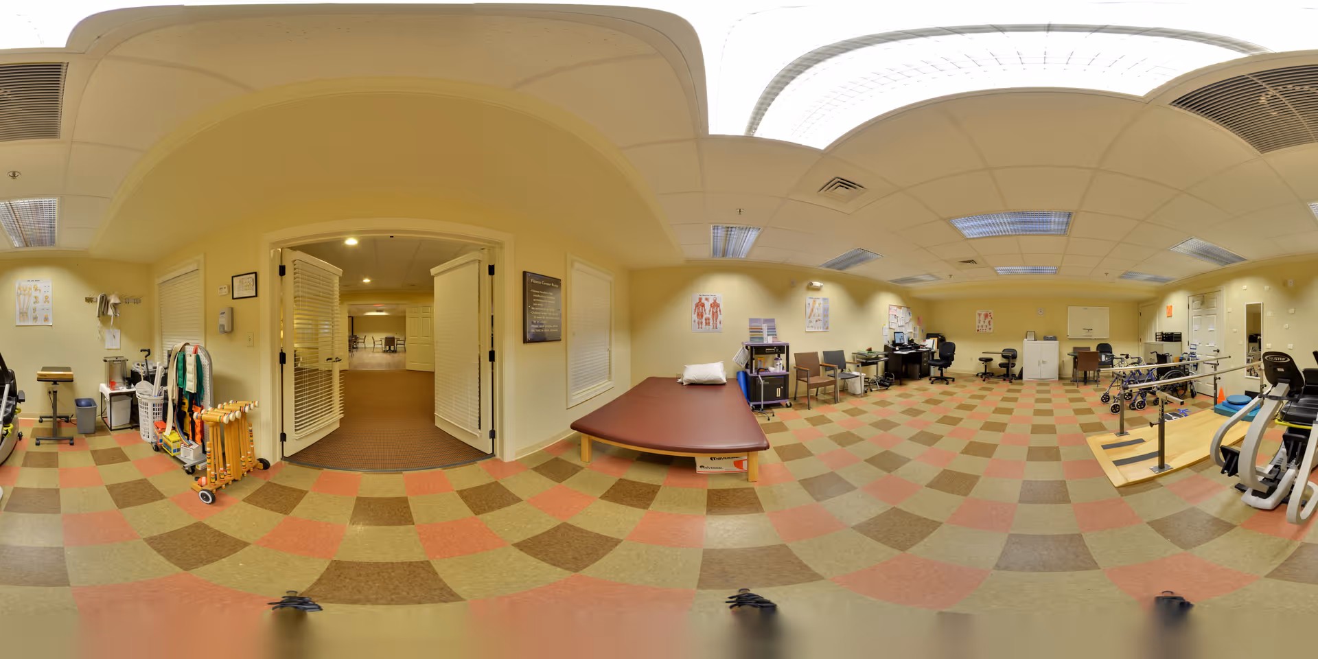 Spacious senior rehab/therapy room with a treatment table, chairs, exercise bikes, parallel bars and mobility aids on a checkered floor.
