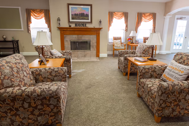 A cozy living room area with floral patterned armchairs arranged around wooden side tables with lamps. There is a fireplace with a wooden mantel decorated with a painting and the word 'HOME'. The room has beige walls, carpeted floor, and windows with orange curtains letting in natural light.