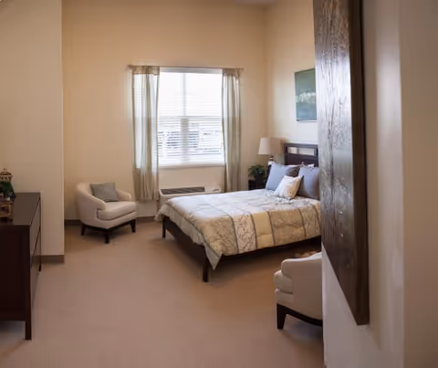 A cozy bedroom with a neatly made bed featuring patterned bedding and multiple pillows. There are two beige armchairs, one near the window and one near the foreground. A window with light curtains lets in natural light. A wooden dresser is on the left side of the room, and a lamp and plant are on a nightstand next to the bed. A large piece of artwork is partially visible on the right wall.
