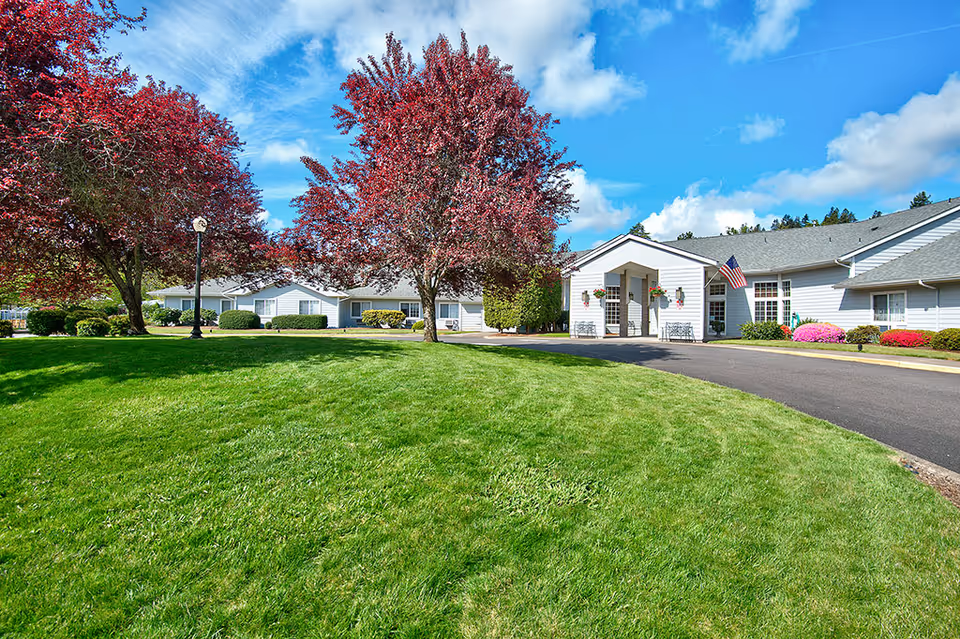 Exterior view of Emerald Valley Assisted Living facility with a well-maintained green lawn, red-leaved trees, a paved driveway, and a white building with an American flag near the entrance under a partly cloudy blue sky.