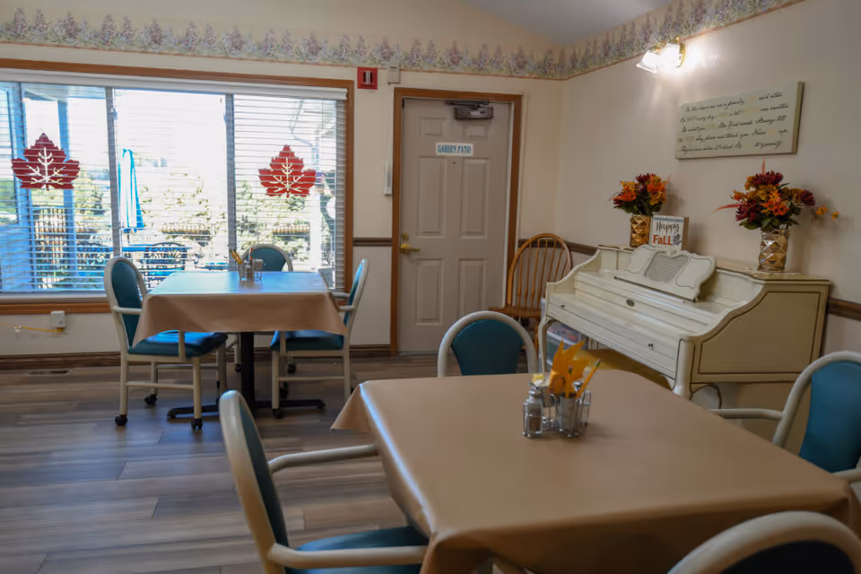 A cozy dining room with tables covered in beige tablecloths and teal chairs. There is a white piano against the wall decorated with two vases of fall-themed flowers and a small sign that says 'Happy Fall.' A door labeled 'Garden Patio' is visible, along with large windows featuring red maple leaf decals. The room has wooden flooring and floral wallpaper border near the ceiling.