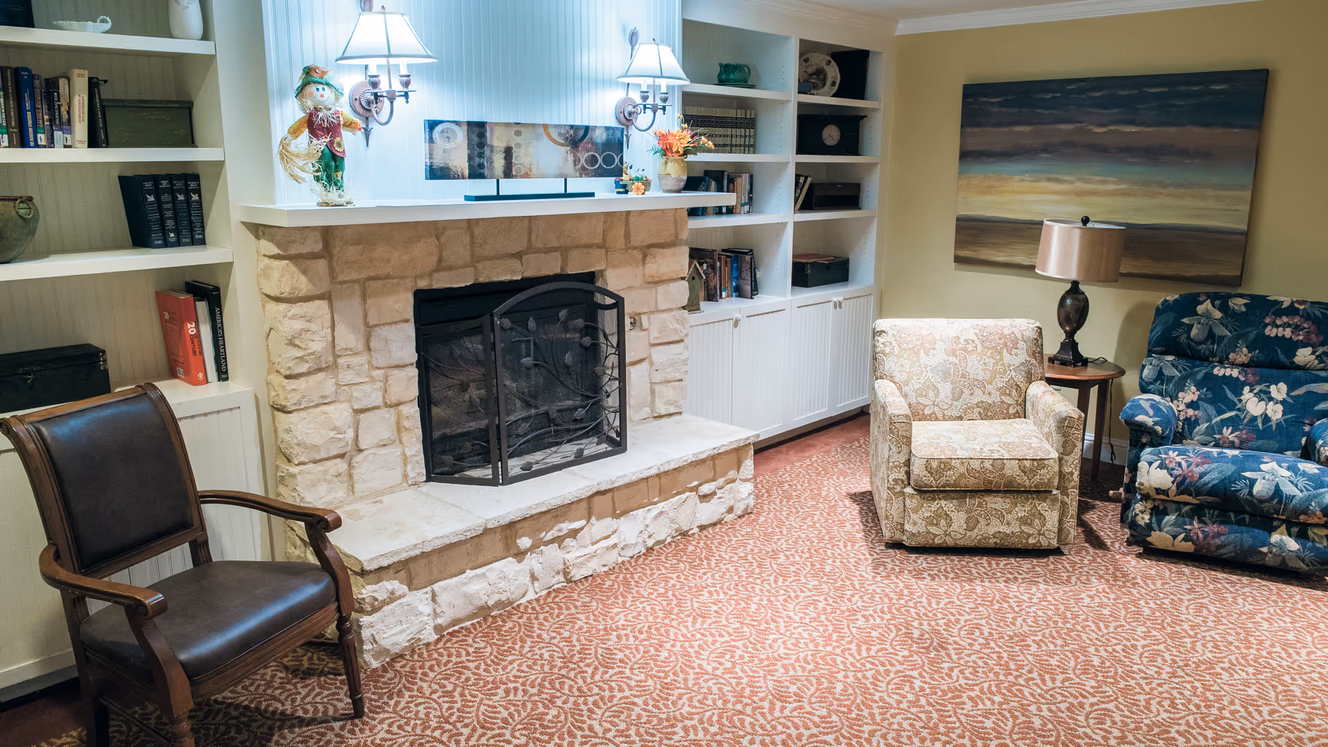 Cozy common room with a stone fireplace, built-in bookshelves, upholstered chairs, a lamp, and patterned carpet.
