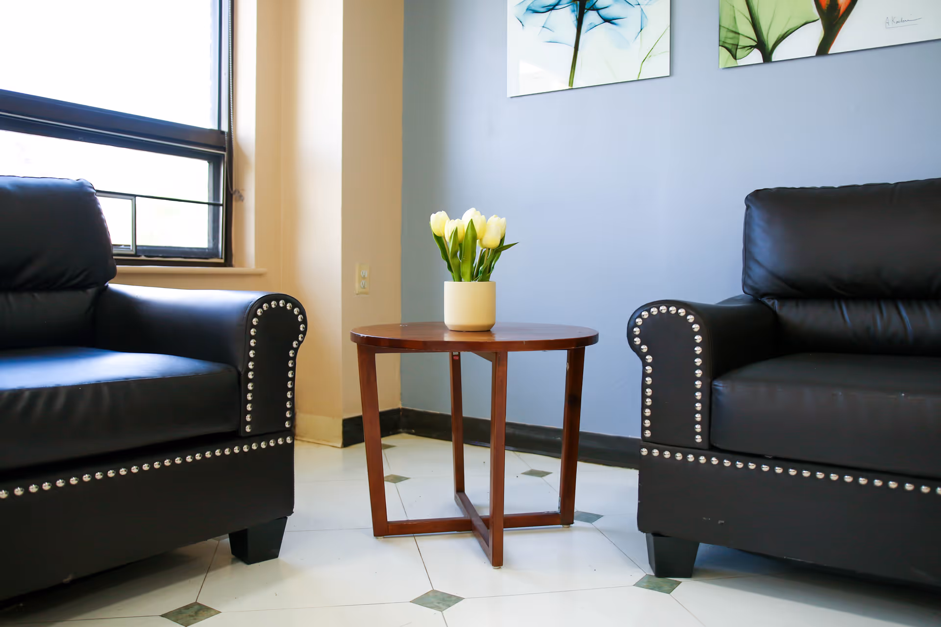 A small seating area with two black leather armchairs featuring silver studs along the arms and base. Between the chairs is a round wooden table with a white vase holding yellow tulips. The room has a window on the left side and light blue walls with two framed floral artworks hanging above the table.