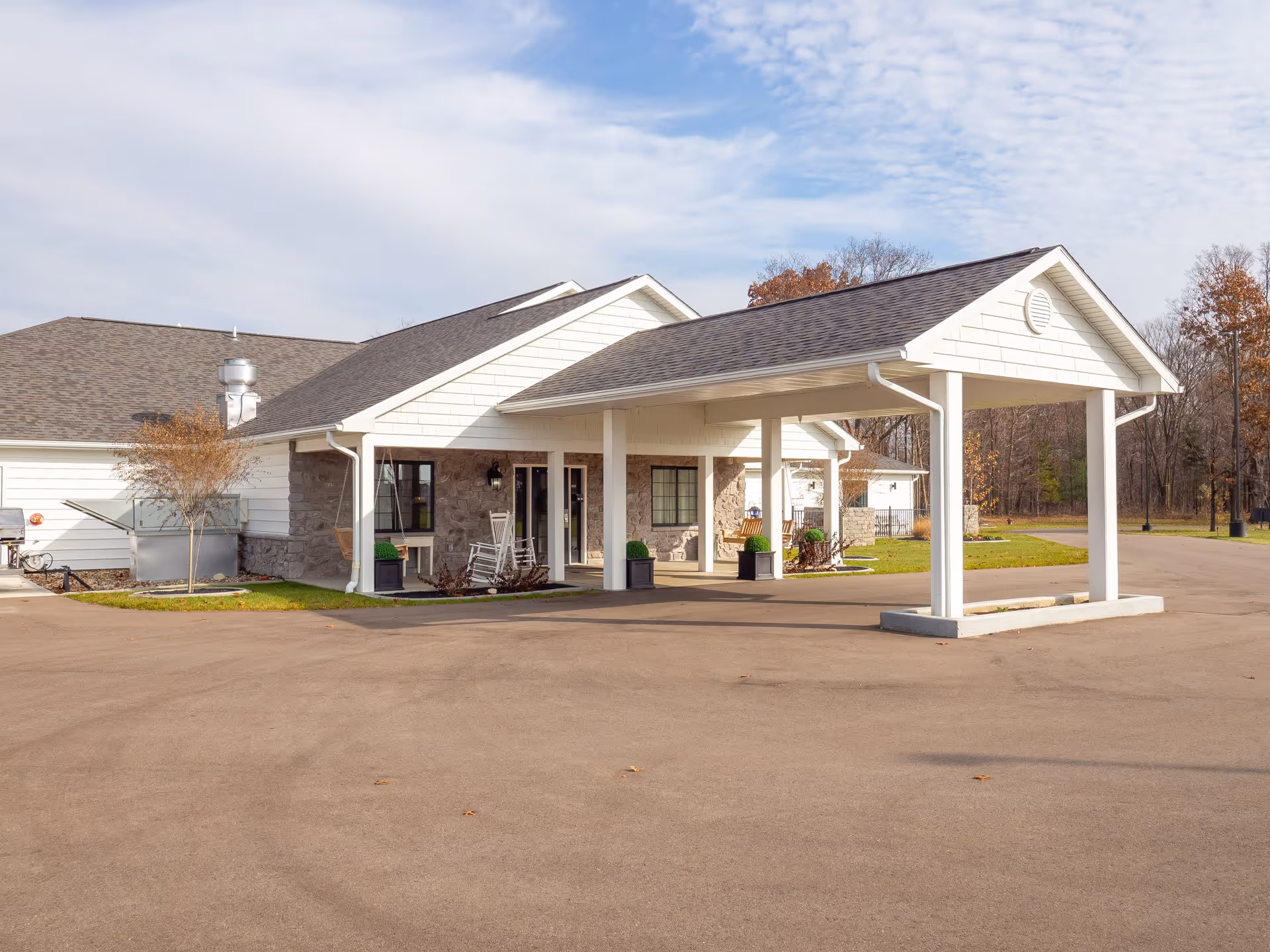 Exterior view of a single-story senior care facility building with a covered entrance supported by white columns. The building has white siding and stone accents, with a paved driveway in front and some trees in the background under a partly cloudy sky.
