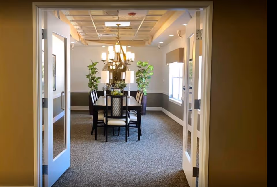 Through open double doors, a dining room with a long table, chairs, chandelier, and potted plants.