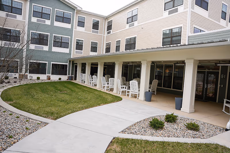 Courtyard with a covered patio lined with white rocking chairs, pathways, and a grassy area in front of a multi-story residential building.