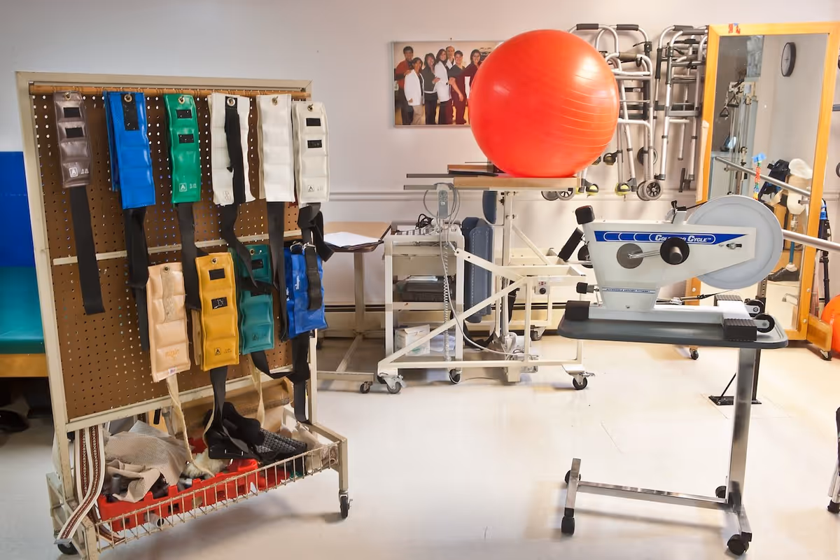 A physical therapy or rehabilitation room with various exercise equipment including a stationary exercise bike, a large red exercise ball on a stand, and a pegboard holding multiple weighted straps. Walkers are hanging on the wall next to a large mirror, and a framed photo of a group of people is mounted on the wall.