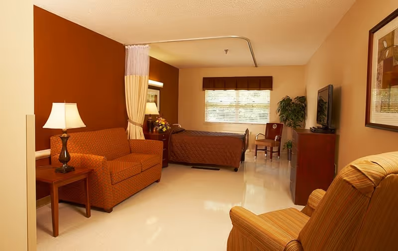 A furnished senior living bedroom with a bed, sofa, armchair, TV, and warm-toned decor.