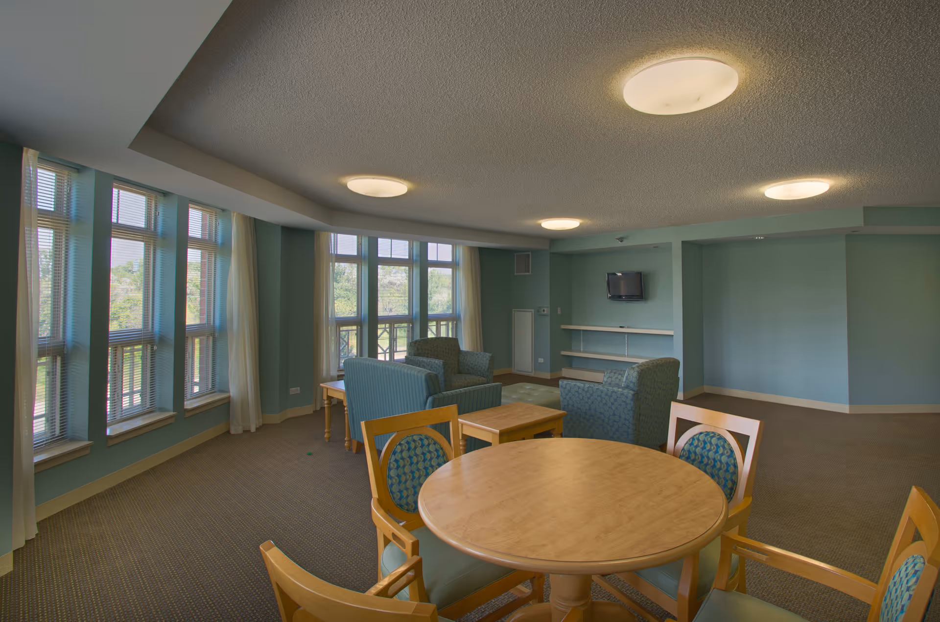 A spacious common area in a senior living facility with large windows letting in natural light. The room features a round wooden table with four chairs in the foreground, and several upholstered armchairs arranged around a small wooden coffee table. The walls are painted light blue, and there is a small wall-mounted TV with shelves beneath it. The ceiling has multiple round light fixtures.