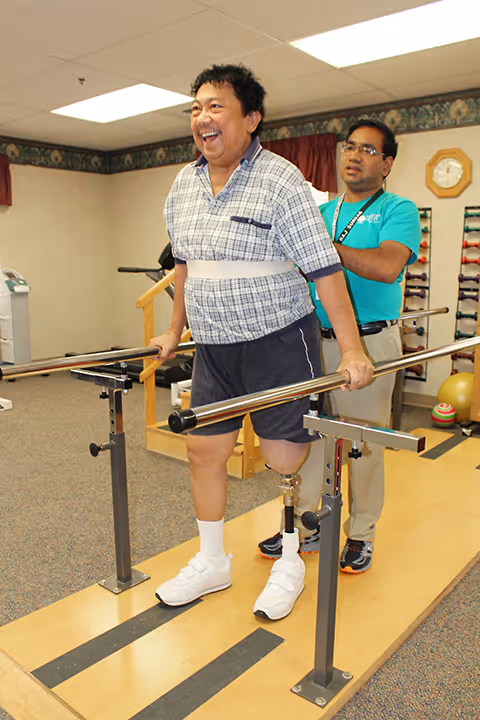 A man with a prosthetic leg is walking with the support of parallel bars in a rehabilitation or physical therapy room, assisted by a caregiver standing behind him. The man is smiling and appears to be making progress in his therapy.