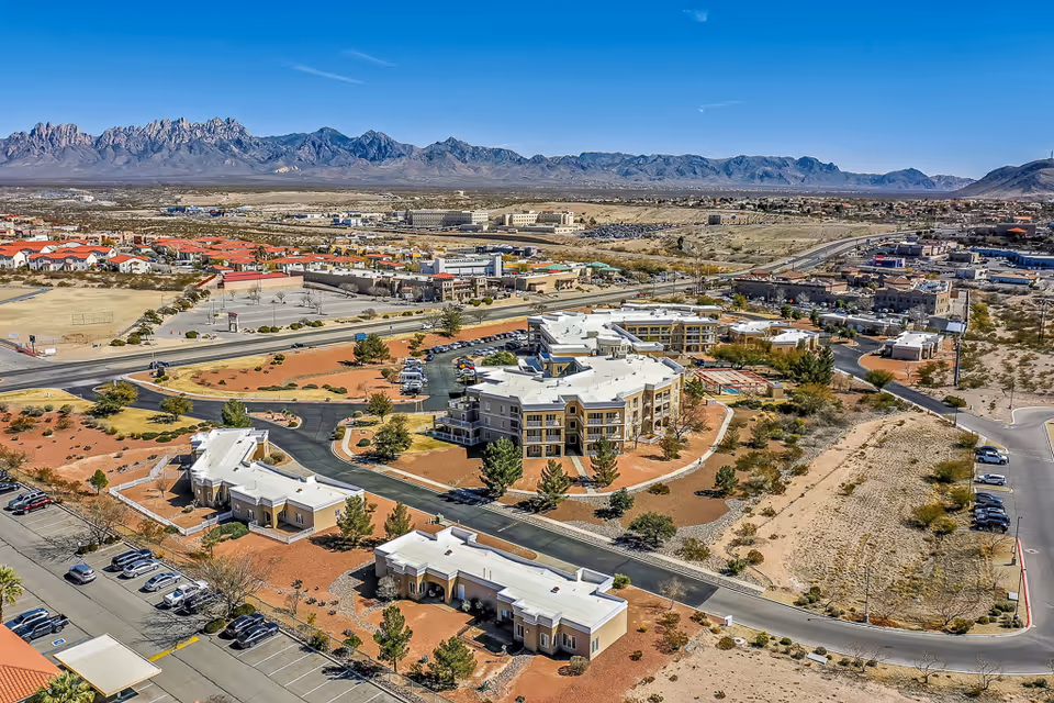 Aerial view of a senior living campus with multiple tan buildings, parking lots, roads, and desert landscaping backed by distant mountains.