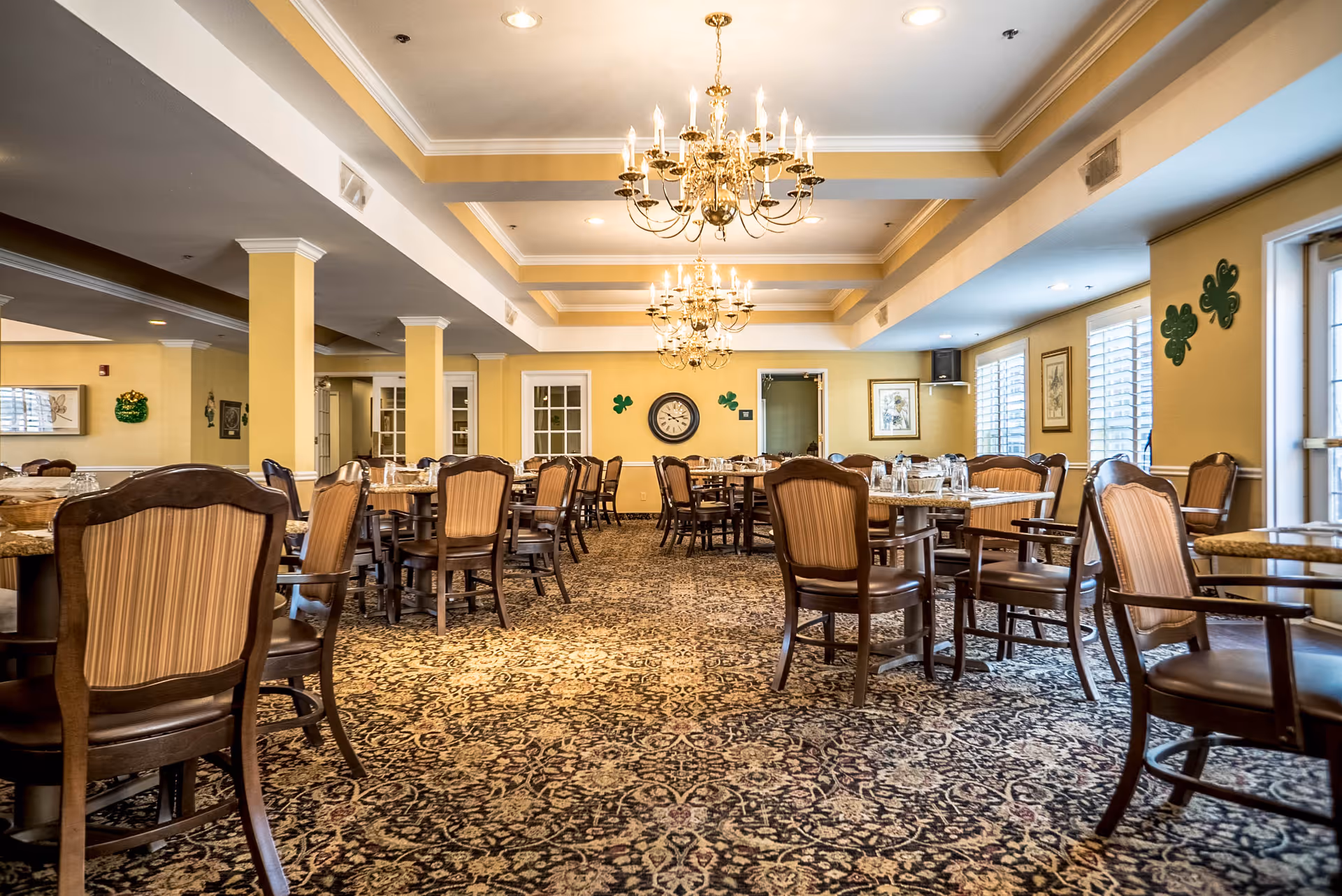 A spacious dining room with multiple wooden tables and chairs arranged neatly. The room features patterned carpet flooring, yellow walls, and several chandeliers hanging from a coffered ceiling. There are windows with white blinds on the right side and decorative wall art including a clock and shamrock decorations.