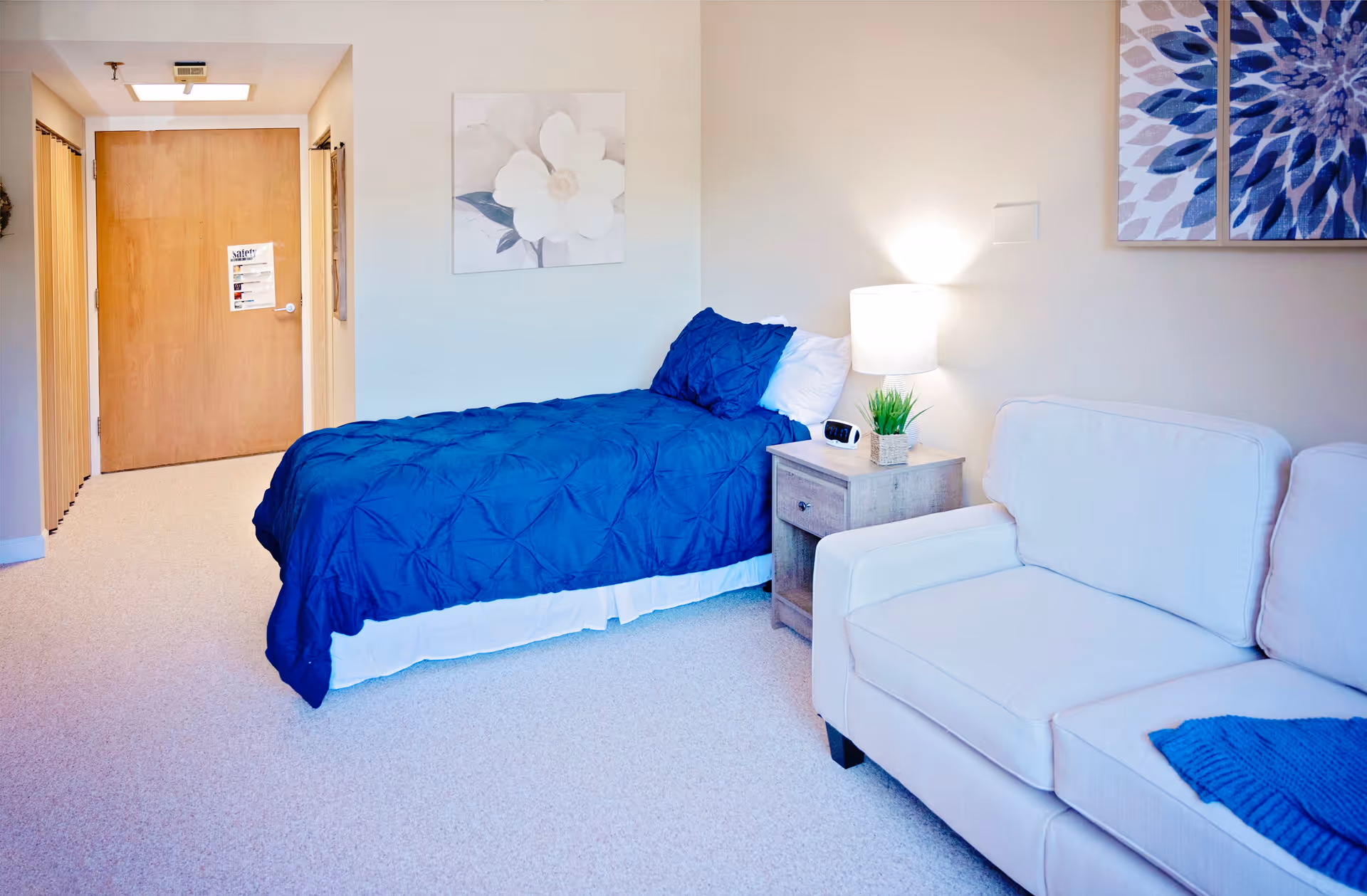 A bright and clean bedroom featuring a single bed with a blue comforter and white sheets, a wooden nightstand with a lamp, a small plant, and an alarm clock. Next to the nightstand is a white cushioned armchair with a blue throw blanket. The walls are decorated with floral artwork, and a wooden door is visible in the background.