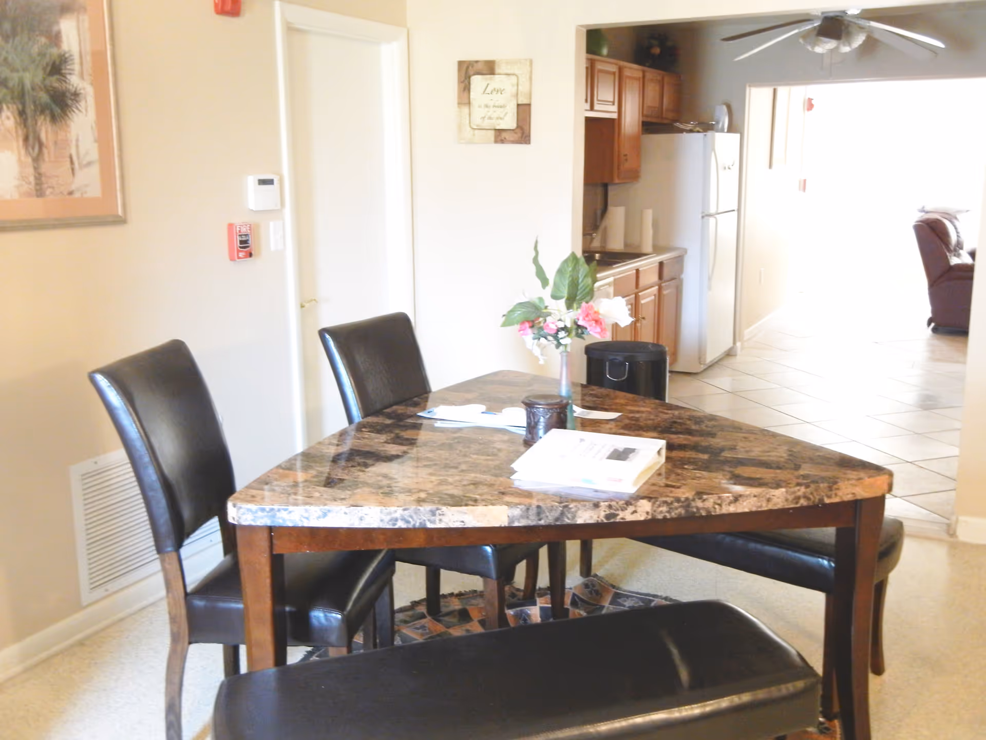 Interior view of a dining area with a triangular marble-top table surrounded by three black leather chairs and a black bench. A vase with pink and white flowers is on the table along with some papers. In the background, there is a small kitchen area with wooden cabinets, a refrigerator, and a trash can. A ceiling fan is visible above, and a living room with a brown recliner chair is seen through an open doorway.