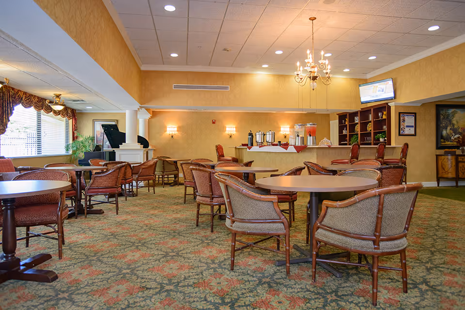 Spacious senior facility dining/lounge area with round tables, upholstered chairs, a beverage buffet and chandelier.