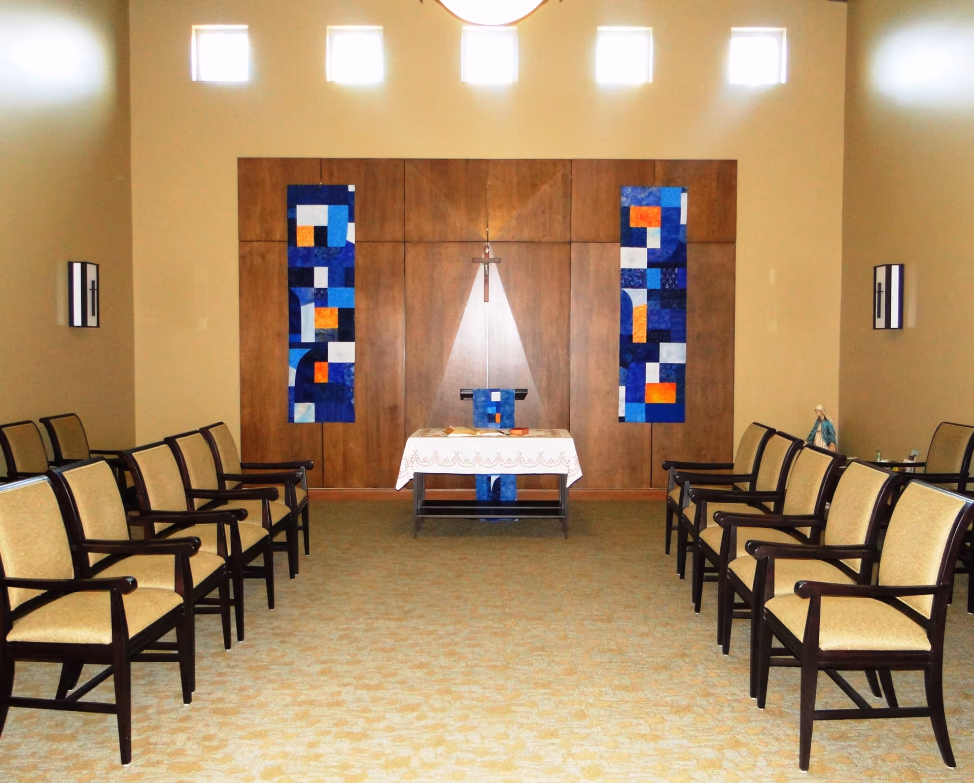 A small chapel or prayer room with two rows of beige cushioned chairs with dark wooden frames facing a wooden altar. The altar is covered with a white cloth and has a cross mounted on the wooden panel behind it. Two vertical blue and orange abstract banners hang on either side of the cross. Small square windows near the ceiling let in natural light.