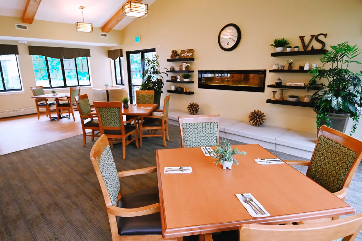 A bright dining room in a senior living facility with wooden tables and chairs arranged neatly. Each table has a small plant centerpiece and place settings with napkins and cutlery. The room features large windows with dark valances, a wall-mounted modern fireplace, decorative shelves with plants and ornaments, and a clock on the wall. There are also ceiling lights and a large potted plant near the entrance door.