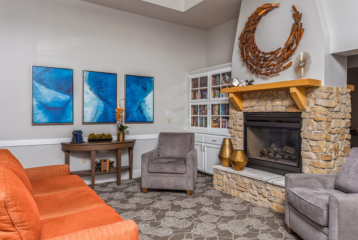 A cozy living room area featuring an orange sofa, two gray armchairs, a stone fireplace with a wooden mantel, and decorative items including a large metal crescent wall art above the fireplace. There is a wooden console table with decorative items and three blue abstract paintings on the wall. A white built-in bookshelf filled with books is next to the fireplace.