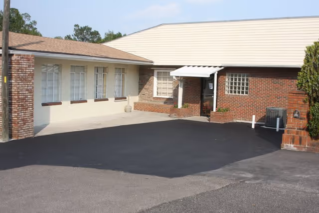 Exterior view of a single-story building with a combination of white and red brick walls, several windows, a small awning over the entrance door, and a paved driveway in front.