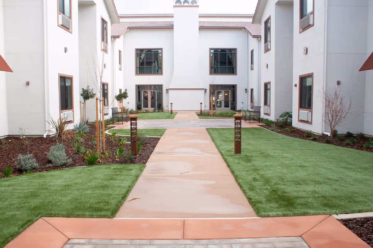A courtyard area at The Villas at Stanford Ranch featuring a concrete walkway flanked by green grass and landscaped garden beds with small trees and shrubs. The courtyard is surrounded by a two-story white building with multiple windows and doors, and there are benches along the sides.