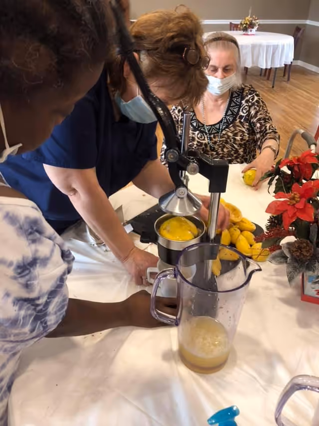 Three women wearing face masks are gathered around a table using a manual citrus juicer to squeeze juice from yellow mangoes. One woman is operating the juicer, while another holds a mango, and the third watches closely. A pitcher with freshly squeezed juice is placed on the table, which is covered with a white tablecloth. In the background, there is a round table with a white tablecloth and a floral centerpiece.
